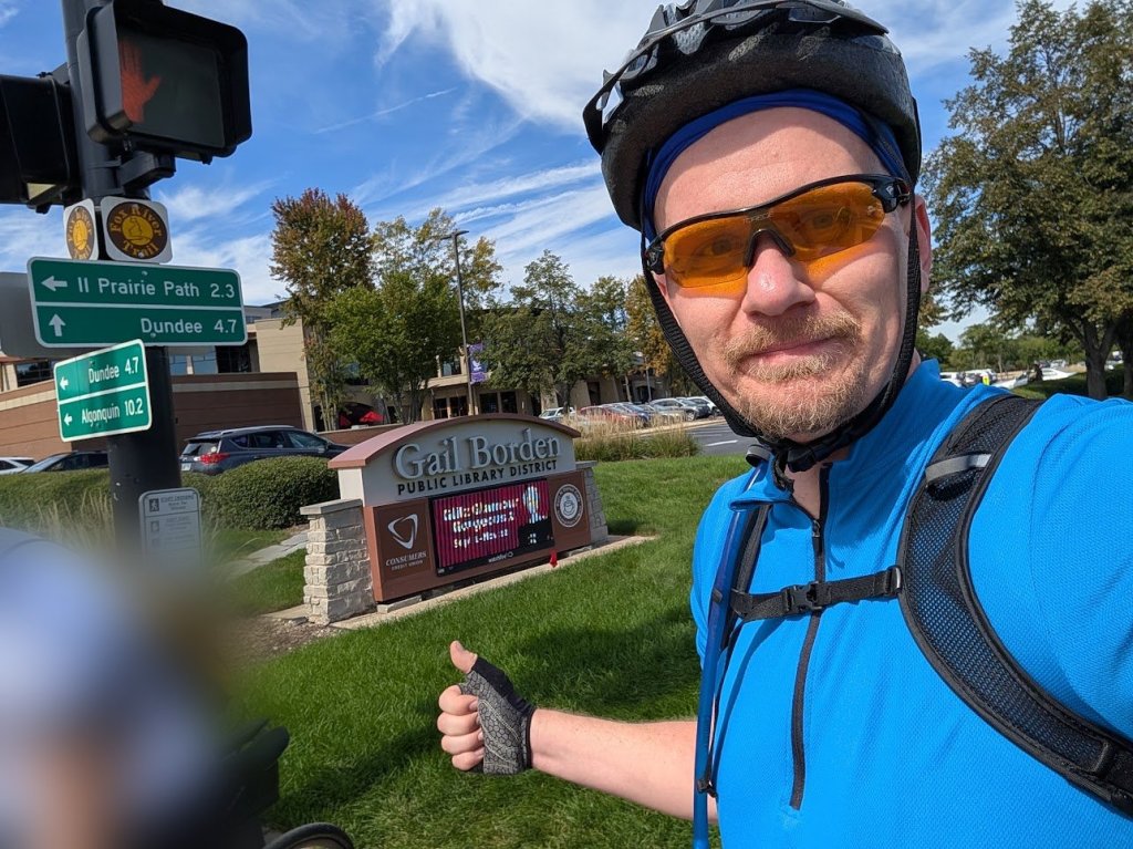 Nick in a blue cycling jersey, helmet, and orange sunglasses giving a thumbs-up next to Fox River Trail signs pointing to the Illinois Prairie Path, Dundee, and Algonquin.