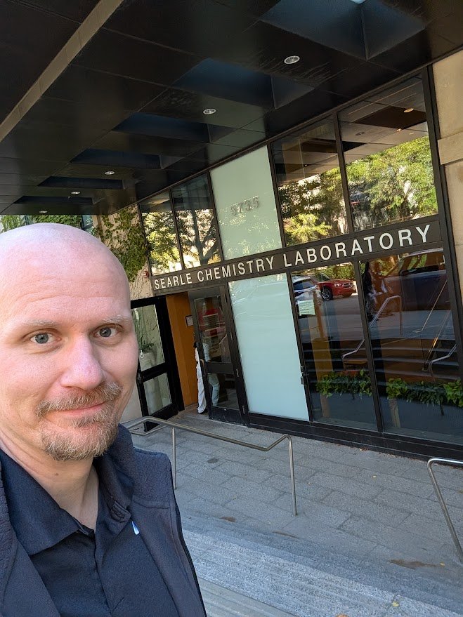Nick Zorn taking a selfie outside the modern glass entrance of the Searle Chemistry Laboratory at the University of Chicago.